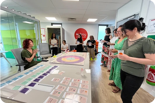 Atelier collaboratif en entreprise : un groupe de personnes participe à une session interactive autour d’un jeu de cartes posé sur une table, avec une animatrice à gauche. L’ambiance est détendue et professionnelle, dans un espace de coworking moderne aux couleurs vives.