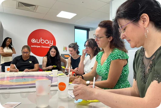 Atelier collaboratif chez Aubay : plusieurs participantes concentrées autour d’une table, écrivant sur des post-its ou manipulant des cartes de jeu colorées, dans une salle lumineuse avec le logo Aubay en fond.